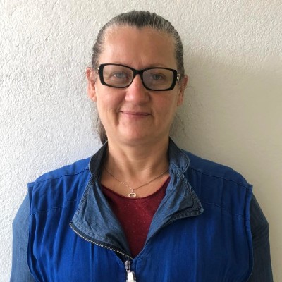 Foto de Eunice Carvalho.  Diante de uma parede branca, Eunice sorri discretamente. Ela tem os cabelos grisalhos presos, usa óculos de armação escura e está com o uniforme azul das equipes de Saúde